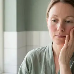 Frau mit geröteten Wangen, die sich sanft ins Gesicht fasst. Sie sucht nach einer Lösung für ihre Rosacea, vielleicht mit Sonnenschutz.