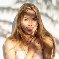 Strahlendes Sommer Make-up: Eine junge Frau mit langen blonden Haaren und grünen Augen lächelt in die Kamera, während Sonnenlicht durch Schatten fällt.