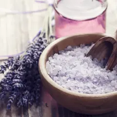Lavendel und Kristallsalz in einer Holzschale, ideal zum badesalz selber machen. Daneben eine kleine Flasche mit lila Flüssigkeit.