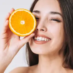 Strahlendes Lächeln mit einer Orangenscheibe vor dem Auge. Vitamin C kann bei Rosacea helfen, die Haut zu beruhigen und zu regenerieren.