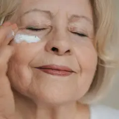 Eine ältere Frau mit blonden Haaren trägt eine Augencreme auf. Ihre welke Haut zeigt die Spuren der Zeit, aber sie pflegt sie liebevoll.