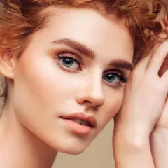 Ein Porträt einer jungen Frau mit roten Haaren und Sommersprossen. Ihre **blauen Augen betonen** ihre natürliche Schönheit, während ihre Hände sanft ihr Gesicht umrahmen.