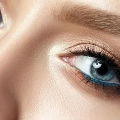 Nahaufnahme eines Gesichts mit makelloser Haut und dramatischem Make-up. Die blau grauen Augen sind mit Lidschatten in Bronze- und Goldtönen sowie einem leuchtend blauen Eyeliner betont.