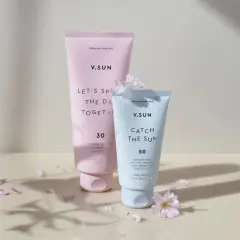 Zwei V.SUN Sonnencreme-Tuben: eine rosafarbene mit LSF 30 und eine hellblaue mit LSF 50, umgeben von zarten Blütenblättern.