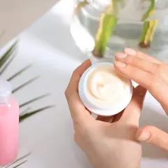 Hände öffnen einen Tiegel mit Creme. Die Creme enthält Urea, das Feuchtigkeit spendet. Daneben steht eine rosa Flasche.