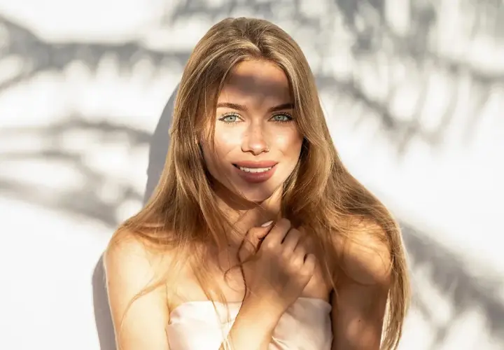 Strahlendes Sommer Make-up: Eine junge Frau mit langen blonden Haaren und grünen Augen lächelt in die Kamera, während Sonnenlicht durch Schatten fällt.