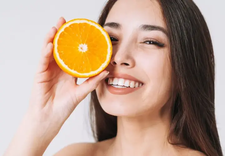 Strahlendes Lächeln mit einer Orangenscheibe vor dem Auge. Vitamin C kann bei Rosacea helfen, die Haut zu beruhigen und zu regenerieren.