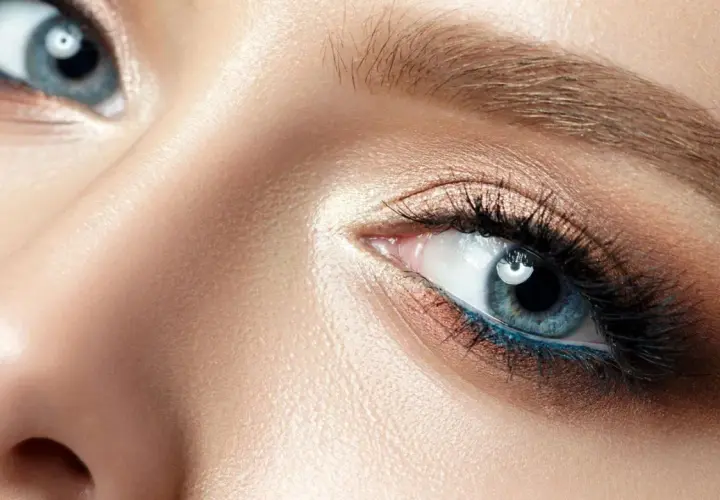 Nahaufnahme eines Gesichts mit makelloser Haut und dramatischem Make-up. Die blau grauen Augen sind mit Lidschatten in Bronze- und Goldtönen sowie einem leuchtend blauen Eyeliner betont.