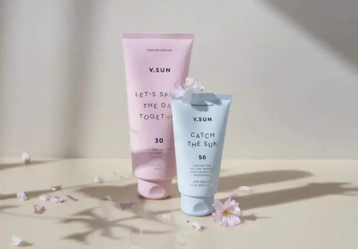 Zwei V.SUN Sonnencreme-Tuben: eine rosafarbene mit LSF 30 und eine hellblaue mit LSF 50, umgeben von zarten Blütenblättern.