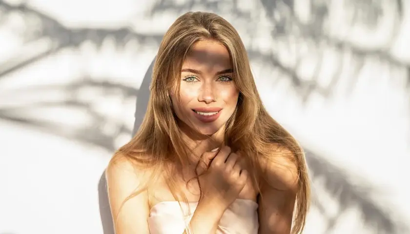 Strahlendes Sommer Make-up: Eine junge Frau mit langen blonden Haaren und grünen Augen lächelt in die Kamera, während Sonnenlicht durch Schatten fällt.