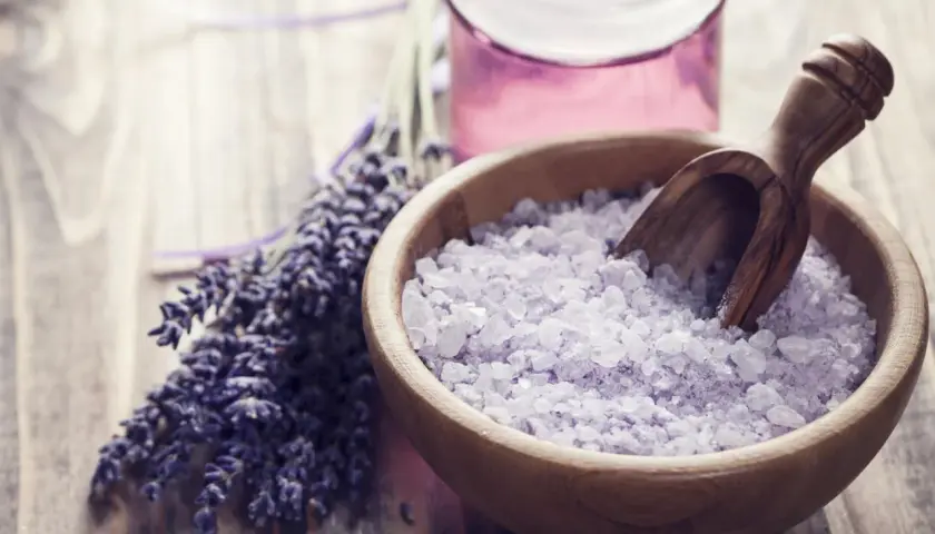 Lavendel und Kristallsalz in einer Holzschale, ideal zum badesalz selber machen. Daneben eine kleine Flasche mit lila Flüssigkeit.