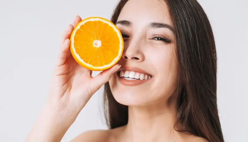 Strahlendes Lächeln mit einer Orangenscheibe vor dem Auge. Vitamin C kann bei Rosacea helfen, die Haut zu beruhigen und zu regenerieren.