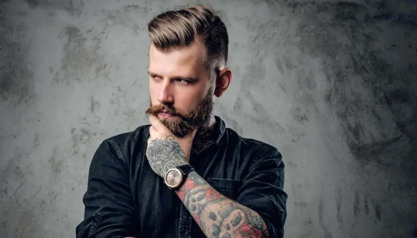 Ein Mann mit Bart und Tattoos, dessen Frisur mit Pomade gestylt ist. Er denkt nach, was ist Pomade?