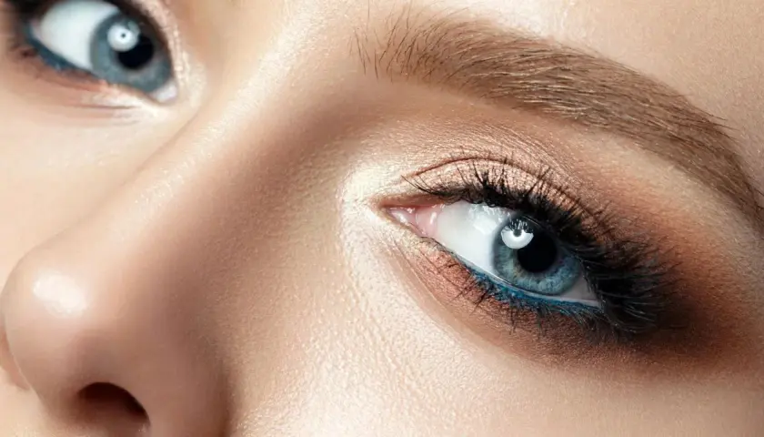 Nahaufnahme eines Gesichts mit makelloser Haut und dramatischem Make-up. Die blau grauen Augen sind mit Lidschatten in Bronze- und Goldtönen sowie einem leuchtend blauen Eyeliner betont.