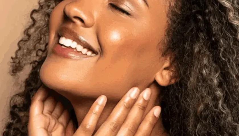 Strahlendes Lächeln einer Frau mit lockigem Haar. Ihre Haut wirkt frisch und glatt, ein Zeichen für gute Pflege und regelmäßige Exfoliation.