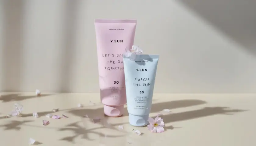Zwei V.SUN Sonnencreme-Tuben: eine rosafarbene mit LSF 30 und eine hellblaue mit LSF 50, umgeben von zarten Blütenblättern.