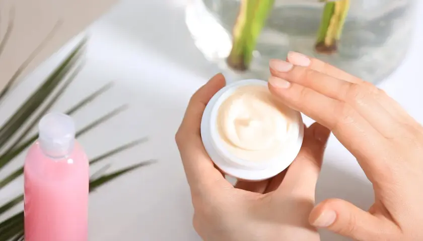 Hände öffnen einen Tiegel mit Creme. Die Creme enthält Urea, das Feuchtigkeit spendet. Daneben steht eine rosa Flasche.