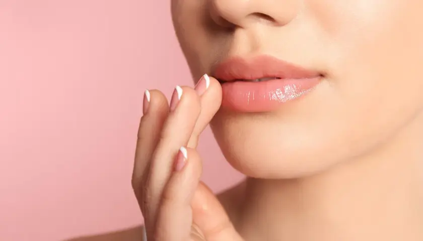 Nahaufnahme von Lippen, die gerade Lipgloss auftragen. Die Finger mit französischem Maniküre-Nageldesign berühren sanft die Lippen.