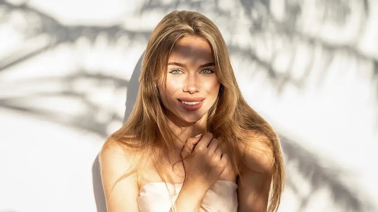 Strahlendes Sommer Make-up: Eine junge Frau mit langen blonden Haaren und grünen Augen lächelt in die Kamera, während Sonnenlicht durch Schatten fällt.