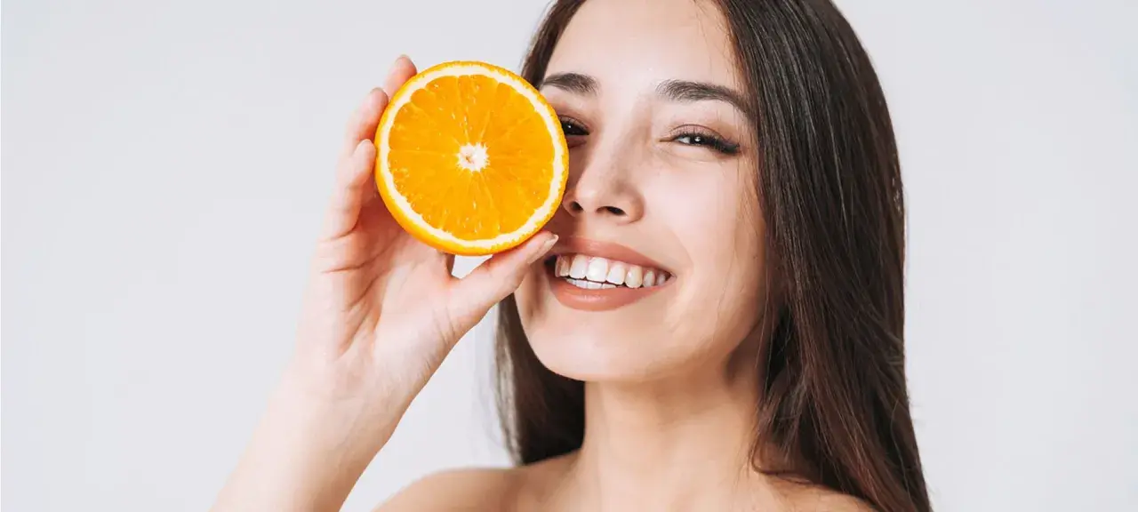Strahlendes Lächeln mit einer Orangenscheibe vor dem Auge. Vitamin C kann bei Rosacea helfen, die Haut zu beruhigen und zu regenerieren.