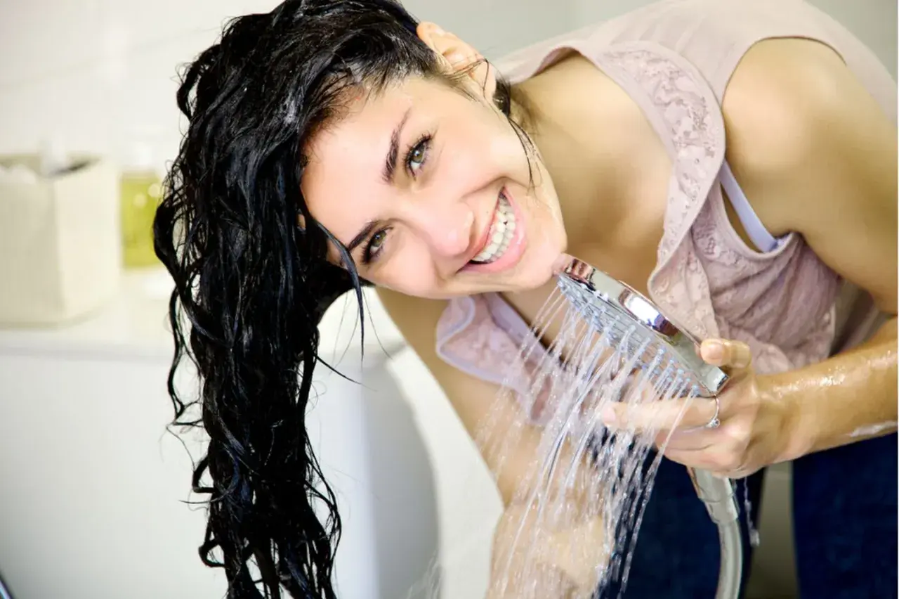 Frau mit nassem Haar lacht unter der Dusche. Sie fragt sich, was sind Silikone im Shampoo und wie sie das Haar glänzend machen.