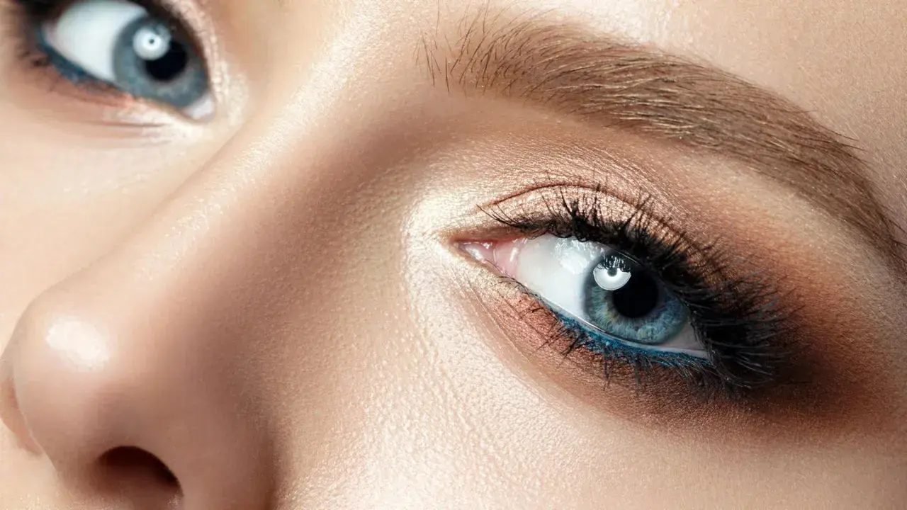 Nahaufnahme eines Gesichts mit makelloser Haut und dramatischem Make-up. Die blau grauen Augen sind mit Lidschatten in Bronze- und Goldtönen sowie einem leuchtend blauen Eyeliner betont.