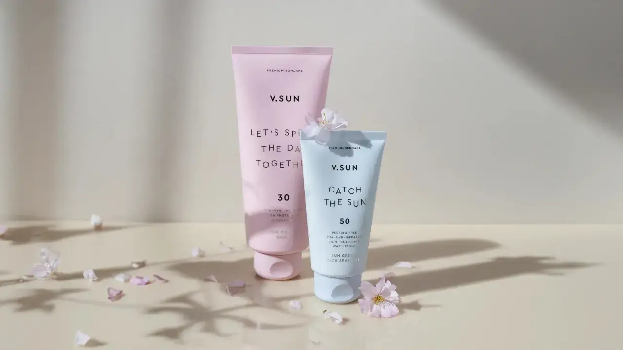 Zwei V.SUN Sonnencreme-Tuben: eine rosafarbene mit LSF 30 und eine hellblaue mit LSF 50, umgeben von zarten Blütenblättern.