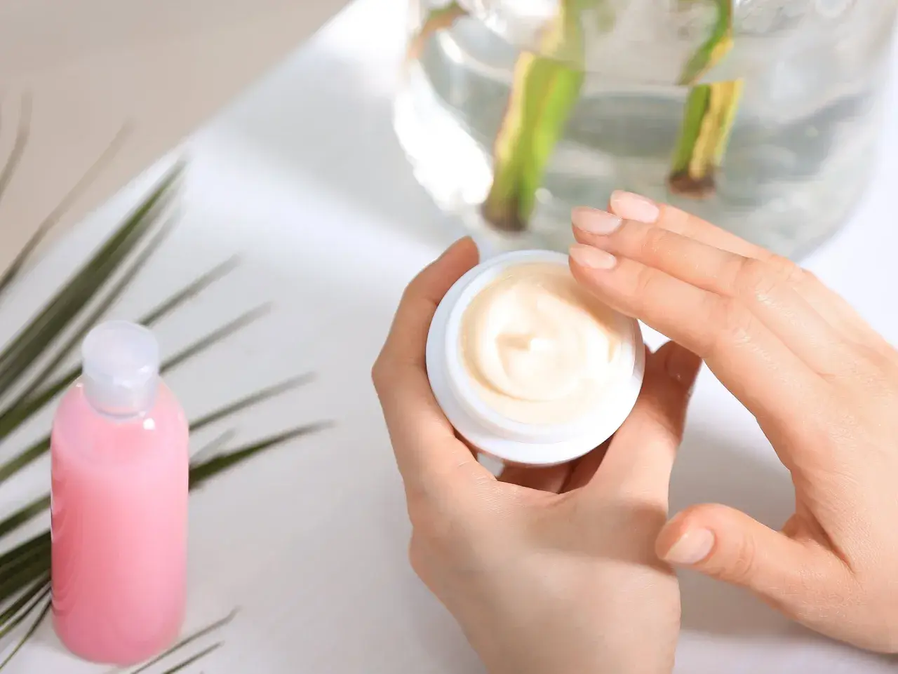 Hände öffnen einen Tiegel mit Creme. Die Creme enthält Urea, das Feuchtigkeit spendet. Daneben steht eine rosa Flasche.