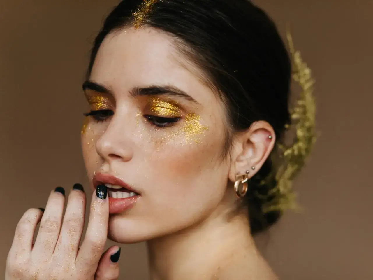 Glitzernder Make-up-Look f&uuml;r Silvester: Goldener Lidschatten, Glitzer im Gesicht und schwarze N&auml;gel.