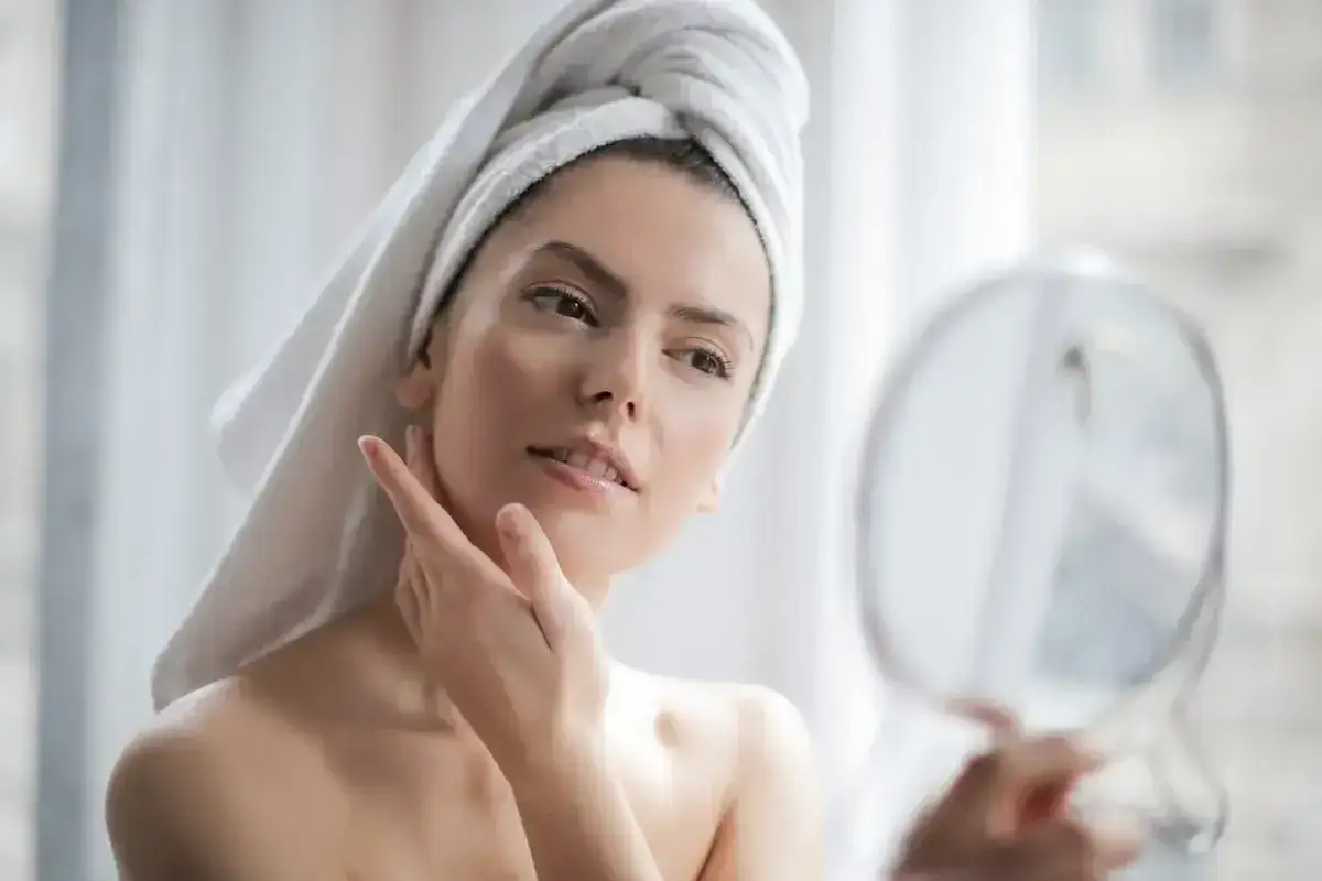 Frau mit Handtuch auf dem Kopf streicht sich &uuml;ber das Gesicht, das eine makellose glass skin zeigt. Sie blickt in einen Spiegel.