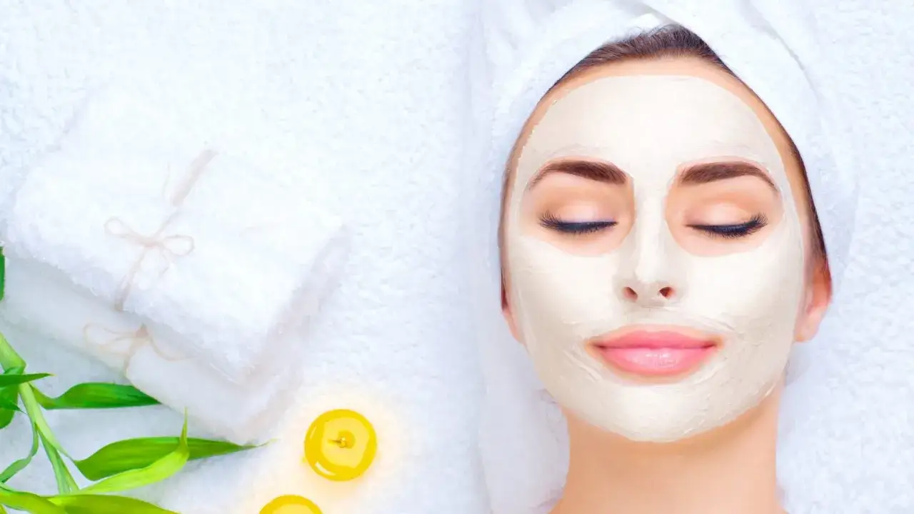 Entspannende Gesichtsmaske morgens oder abends. Eine Frau mit geschlossenen Augen und einer cremigen Maske im Gesicht, umgeben von Handtüchern und Kerzen.
